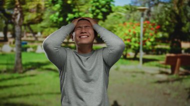Parktaki genç Çinli adam, verimli yeşillikler ve güneşli bir günde park banklarıyla çevrili çeşitli duygular gösteriyor..