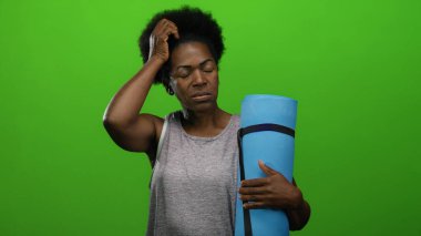 Mavi yoga minderi tutan kadın, yeşil arka plana karşı gergin, afro saç stili, kolsuz bluz giyiyor, sakin ve düşünceli..