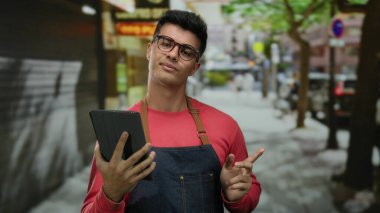 Önlük ve gözlük takmış, güvenli bir şekilde ağaçlarla dışarıda duran ve profesyonel ama gelişigüzel bir şehir havası katan genç bir adam..