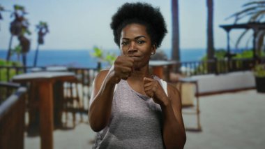 Deniz kenarında güneşli bir restoranın terasında boks yapan bir kadın, manzaralı bir arka planda açık hava eğitimi sırasında kararlılık ve güç sergiliyor..