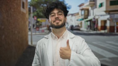 Sakallı, gözlüklü, beyaz önlüklü, güneşli bir şehir sokağında güven ve pozitiflik gösteren genç bir İspanyol..