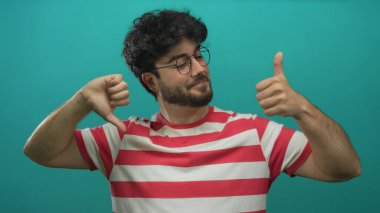 Çizgili tişörtlü, sakallı genç adam yeşil arka plana başparmak işareti yapıyor. Düşünceli bir bakışla karışık tepkiler veriyor..