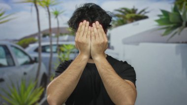 Sakallı genç bir adam güneşli bir günde dışarıda komik suratlar ve jestler yaparak yemyeşil ve açık mavi gökyüzü ile neşeli ve hayat dolu bir hava yaratıyor..