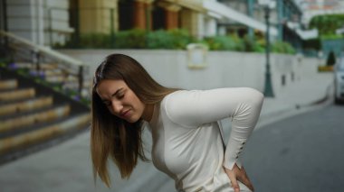 Beyaz kazaklı kadın arka planda merdivenleri olan bir şehir sokağında acı içinde sırtını tutuyor..