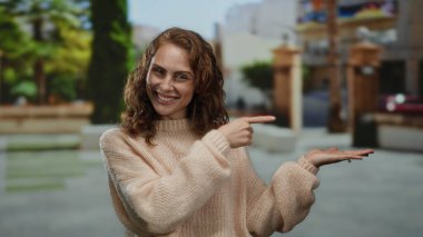 Genç bir kadın, açık hava parkında uzanan avuç içini göstererek gülümsüyor, rahat bir kazak giyiyor, güneşli günün tadını çıkarıyor ve bulanık yeşillik ve kentsel unsurlarla çevrili..