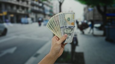 Bir şehir caddesinde dolarları elinde tutan bir adam, Amerikan banknotlarını sergiliyor, açık havada şehir ortamında.