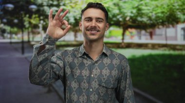 Genç İspanyol adam desenli gömleğiyle gülümsüyor, sakin parkta işaret veriyor yemyeşil ağaçların altında ve sessiz arka planda, rahat açık hava atmosferini yakalıyor..