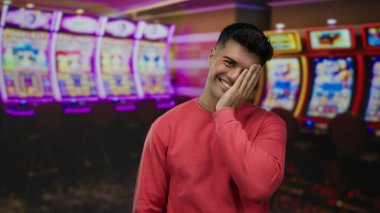 Canlı bir kumarhanede genç bir adam renkli kumar makinelerinin yanında neşeli bir ifade ve rahat bir atmosfer sergileyerek durur..