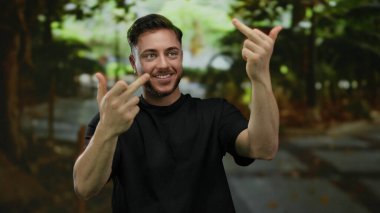 Parkta sakallı genç adam iki orta parmağı havada, etrafı yeşilliklerle çevrili, siyah gömlekli, dışarıda duygularını ifade eden kaba bir jest yapıyor..