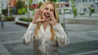 Kalabalık bir şehir caddesinde neşeli bir şekilde bağıran, beyaz bir gömlek giyen, sarı saçlı, canlı ve canlı bir şehir manzarası çeken bir kadın..