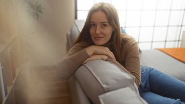 Kadın rahat bir kanepede rahat bir şekilde dinleniyor. İçerde yumuşak bir gülümseme, sakin ve rahat bir ev atmosferiyle çevrili..