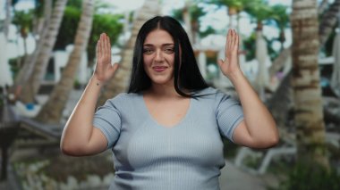 Tatil köyündeki kadın, canlı bir atmosferi olan palmiye ağaçlarının altında neşeli bir şekilde poz verirken el hareketleri yapıyor..