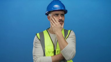 Yansıtıcı yelek ve kasketli genç İspanyol adam, izole edilmiş mavi duvar arkaplanına karşı düşünceli bir şekilde duruyor. İnşaatı ve planlamayı simgeliyor..
