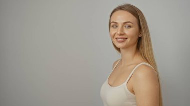 Beyaz bir arka plana güvenle gülümseyen bir kadın pozitif gençlik enerjisi ve güzelliğinin ifadesi ile sarı saçlarını ve kolsuz bluzunu gösteriyor..