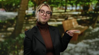 Bir kadın gülümsüyor ve açık hava parkında gözlük takıyor ve siyah bir ceket giyiyor yemyeşil yapraklar ve gün ışığında ahşap bir banka yaslanıyor..