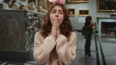 Tavşan kulağı takan genç bir kadın, sanat eserleriyle çevrili bir galeri ortamında duygularını ifade ediyor, yaratıcı bir ortamda eğlence ve merak taşıyor..