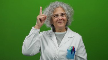 Beyaz laboratuvar önlüklü, yukarı doğru kıvırcık gri saçlı, parlak yeşil duvar arkaplanına karşı izole edilmiş bir kadın, bir fikri ya da ilham anını tasvir ediyor..