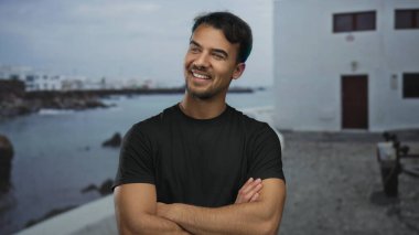 Genç, İspanyol asıllı bir adam, kolları bağlı, güvenli bir şekilde deniz kenarındaki gezinti alanında gülümsüyor, sakin bir okyanus arka planı, dış mekanı güzelleştiriyor..