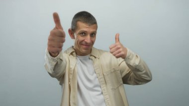 Gülümseyen bir adam beyaz arka planda yalnız, bej gömlek ve kısa saçlı beyaz tişört giyiyor ve pozitif jest yapıyor.