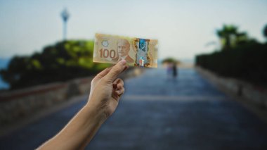 Kanadalı 100 dolarlık banknotu açık şehir ortamında bir sokakta tutarak, odak noktasında bir adamla gelişigüzel bir şehir anını yakalıyor..