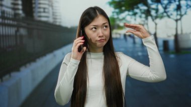 Şehir sokağında telefonla dalga geçen, günlük kıyafetler giyen, uzun saçlı, şehir yolunda esprili bir şekilde el kol hareketi yapan, oyuncu ifadeler sergileyen, açık hava sahnesi olan bir kadın..