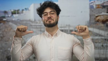 Sakallı, gözlüklü, gülümseyen ve bir inşaat alanında kendini gösteren genç bir adam, günlük kıyafetlere olan güvenini dile getiriyor..