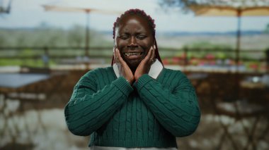 Terasta üzgün bir kadın, elleri yüzün üzerinde yeşil kazak giyiyor. Restoranın arka planında güneşli bir günde bulanık sandalyeler ve ağaçlar var.