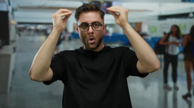 Kapalı bir havaalanı terminalinde gözlük takan sakallı genç adam hareketli bir ortamda gezginlerle çevrili modern bir seyahat sahnesi yaratıyor..