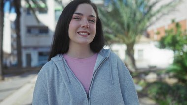 Güneşli bir sokakta palmiye ağaçlarıyla gülümseyen bir kadın, açık havada ellerle kalp oluşturan, sıradan bir kapüşonlu giyen bir kadın..