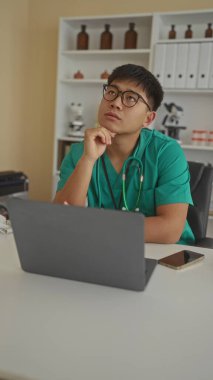 Steteskoplu yeşil önlüklü genç doktor, tıbbi ekipman ve raflarla çevrili modern bir klinikte dizüstü bilgisayar üzerinde çalışırken düşünceli görünüyor..