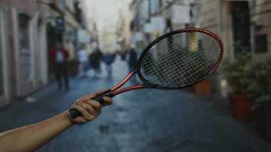 Yoğun bir şehir caddesinde tenis raketi tutan adam, sokak sporlarını şehir zeminine karşı sergiliyor, atletik aktiviteleri günlük yaşam ve mimari ile harmanlıyor..