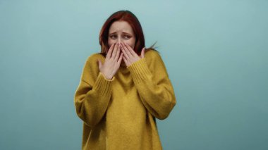 Sarı kazaklı kızıl saçlı bir kadın mavi bir duvara karşı utangaç bir şekilde poz veriyor, stok medyasında yaratıcı kullanım için izole bir ortamda güzellik ve duyguyu vurguluyor..