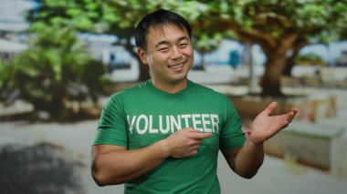 Yeşil gönüllü gömleğiyle gülümseyen genç adam güneşli parkta açık havada, bulanık ağaçlar ve banklarla çevrili, dost canlısı bir toplum atmosferini işaret ediyor..