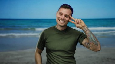Dövmeli gülümseyen adam deniz kenarında barış işareti yapıyor. Arka planda okyanus var. Güneşli gökyüzünün altında kumsal gününün tadını çıkarıyor..