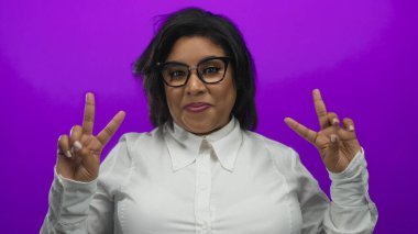 Gözlüklü ve beyaz gömlekli kadın canlı mor arka planda barış işareti yaparken kendine güvenen bir şekilde gülümsüyor.