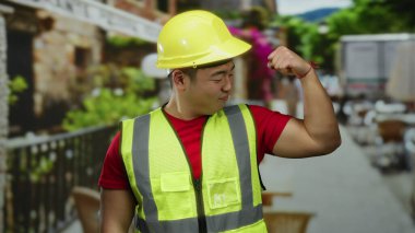 İnşaat malzemesi giymiş bir adam sokak ortasında kaslarını esnetiyor, sarı bir şapka ve yansıtıcı yelek giyerken gücünü ve güvenini gösteriyor..
