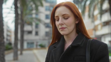 Kızıl saçlı bir kadın, şehir caddesinde palmiye ağaçlarıyla çevrili bir sokakta huzur içinde duruyor. Siyah bir palto giyiyor, şehir hayatının ortasında sakin bir şekilde yaşıyor..