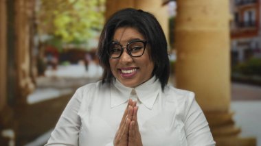 Bir şehir caddesinde el ele gülümseyen bir kadın, gözlük ve beyaz gömlek takıyor, yeşillik ve binaların bulanık arka planına sahip sütunların yanında duruyor..