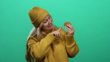Gülümseyen ve kurabiyeye işaret eden sarı kazak giyen ve yeşil arka planda bereli bir kadın.