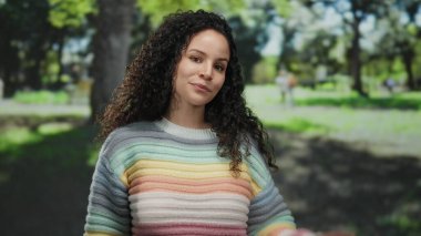 Açık havada güneşli bir günde arka planda ağaçlarla parkın tadını çıkaran renkli kazaklı bir kadın..