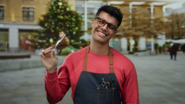 Gözlüklü ve önlüklü İspanyol bir adam arka planda süslü bir Noel ağacı olan şehir sokaklarında makas tutuyor..