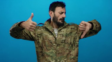 Ordu üniformalı İspanyol bir adam, sakallı, mavi arka planda bir aşağı bir yukarı hareket ediyor, modern asker hayatında karışık duygular ve ikiliği ifade ediyor..