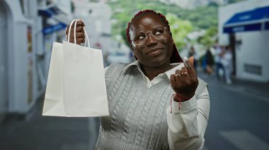 Dışarıda gülümseyen ve parmaklarıyla kalp hareketi yapan bir kadın kalabalık bir şehir sokağında alışveriş torbası tutarken arka planı bulanık şehir atmosferini ve modern hayatın tarzını yakalıyor..