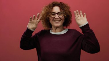 Orta yaşlı, esmer kıvırcık saçlı İspanyol kadın izole edilmiş kırmızı bir arka plan duvarında neşeyle dans ediyor, canlı ifade ve enerji yakalıyor..