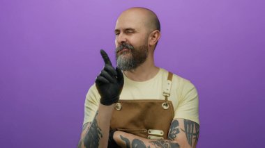 Önlüklü ve siyah eldivenli yakışıklı sakallı adam mor bir arka plana karşı duruyor, düşünceli bir ifade sergiliyor..