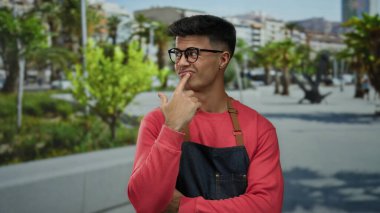 Kırmızı kazaklı ve gözlüklü genç bir adam şehir sokaklarında dikiliyor. Başparmağını kente odaklanmış ifadesiyle işaret ediyor..
