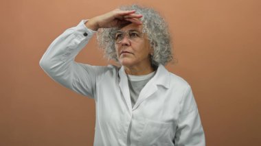 Beyaz önlüklü yaşlı kadın, gri saçlı, kahverengi duvarın karşısında duruyor, düşünceli, izole ve odaklanmış bir şekilde uzaklığa bakıyor..