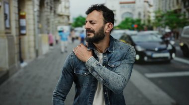 Meşgul bir sokakta kot ceket giyen İspanyol bir adam, dışarıda yansıyor, muhtemelen şehir trafiğinin ortasında omuz ağrısı çekiyor..