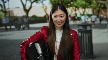 Kırmızı ceketli uzun saçlı kadın, güneşli bir günde ağaçlarla ve arka planda insanlarla şehir sokağında dikilirken miğfer tutuyor..