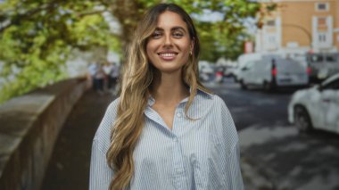 Genç sarışın kadın yüzü, çizgili bluz içinde dalgalı saçlarıyla gülümsüyor yapraklı güneş ışığı kaldırımında; keyif güveni.